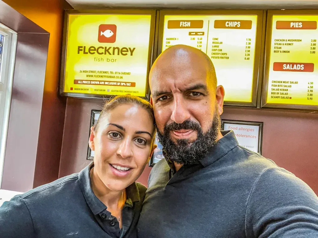 Fleckney Fish Bar signboard displaying menu options including fish, chips, pies, and salads, with two staff members smiling in front of the shop.