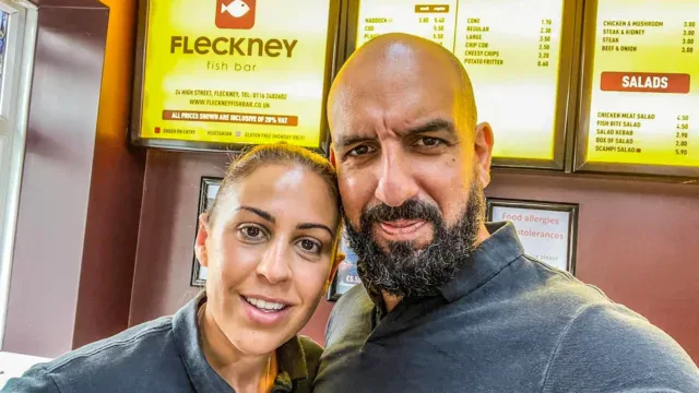 Fleckney Fish Bar signboard displaying menu options including fish, chips, pies, and salads, with two staff members smiling in front of the shop.