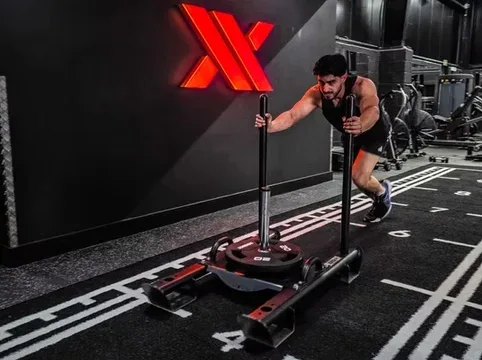 Man pushing a sled at the gym for strength training, with modern gym equipment and a black wall featuring a red logo in the background.