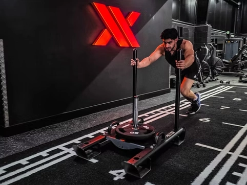 Man pushing a sled at the gym for strength training, with modern gym equipment and a black wall featuring a red logo in the background.