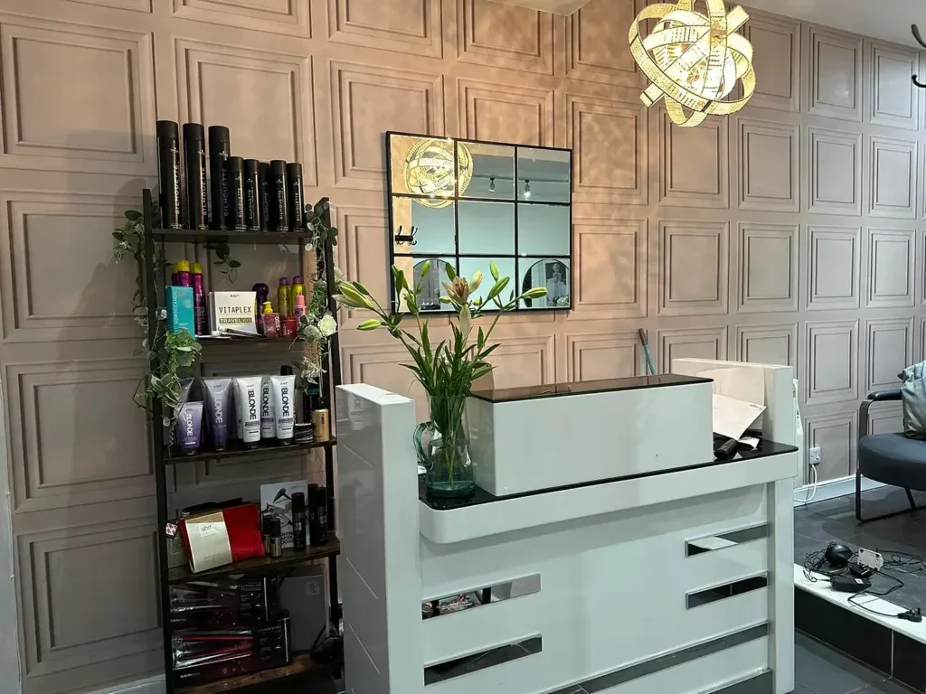 Elegant salon reception with a white counter, decorative wall panels, and a shelf showcasing hair care products.