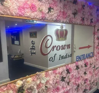 Entrance sign for The Crown of India restaurant, featuring a floral wall with pink and white flowers and a large mirror. Clear arrow indicates the entrance location.