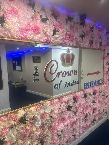 Entrance sign for The Crown of India restaurant, featuring a floral wall with pink and white flowers and a large mirror. Clear arrow indicates the entrance location.