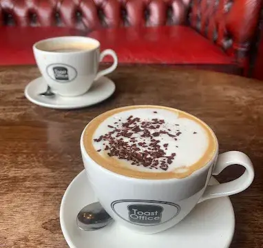 A close-up of two cups of coffee on a wooden table in a cosy cafe setting, featuring a frothy cappuccino with chocolate sprinkles and a smaller espresso in the background.