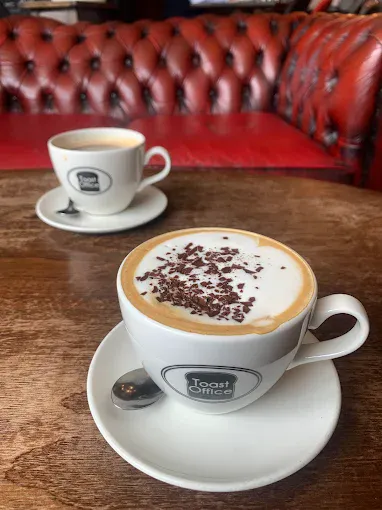 A close-up of two cups of coffee on a wooden table in a cosy cafe setting, featuring a frothy cappuccino with chocolate sprinkles and a smaller espresso in the background.