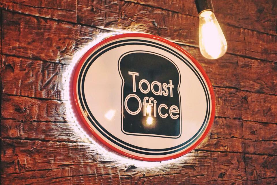 Sign for Toast Office mounted on a rustic brick wall, featuring a retro design with a slice of toast illustration and bold lettering. Perfect for a bakery or café branding.